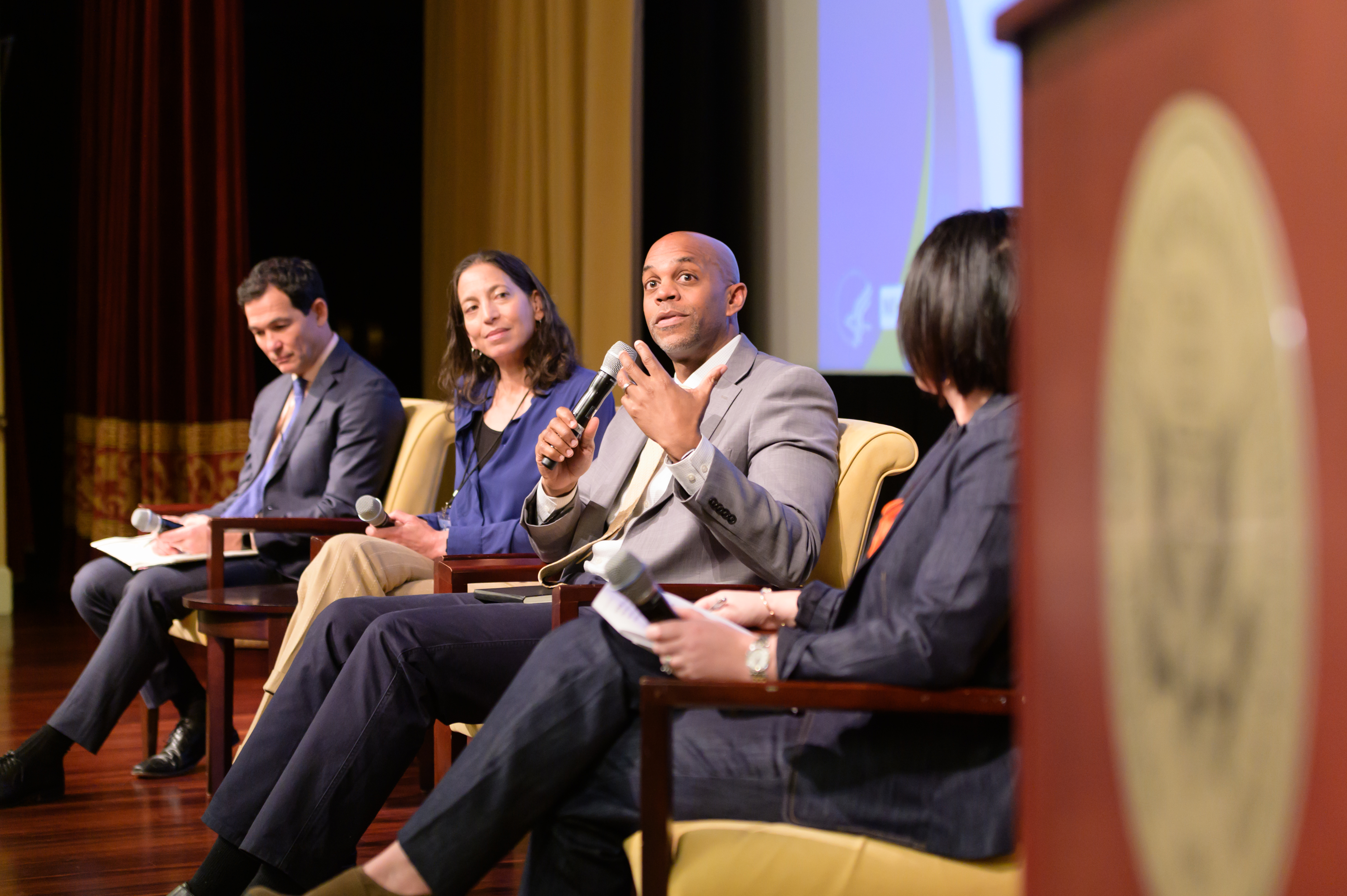 Four people are sitting on a stage in a panel discussion. The person speaking into a microphone is in the center, with three others listening attentively. They are in a formal setting, possibly a conference or seminar.