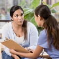 A concerned woman sits and listens intently to a doctor holding a clipboard. They are in a clinic setting with a young child playing with blocks in the background. The focus is on the conversation between the parent and doctor.