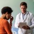 A male provider discusses medical information with a female patient. The provider wears a white lab coat and stethoscope and holds a tablet. The patient sits on the exam table and looks upset or worried, with her hand to her face and pinching the bridge of her nose.