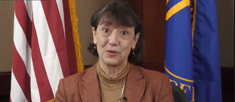 Dr. Monica Bertagnolli shoulder-length dark hair wearing a brown blazer and a tan turtleneck is speaking in front of an American flag and a blue flag.